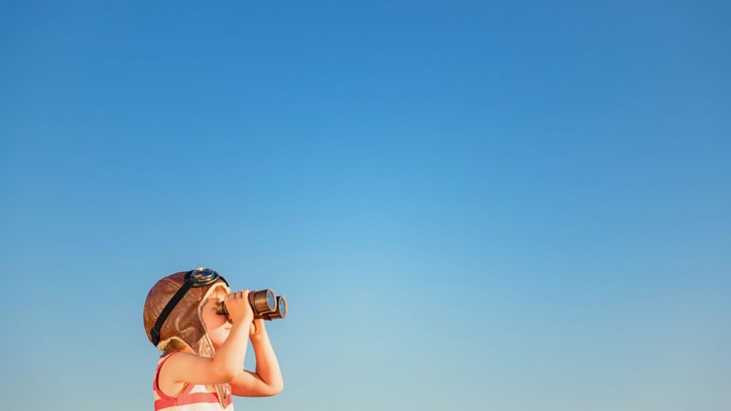 Ein Kind erforscht die Weite mit einem Fernglas vor einem blauen Himmel. Es geht um das Konzept Demokratische Schule.